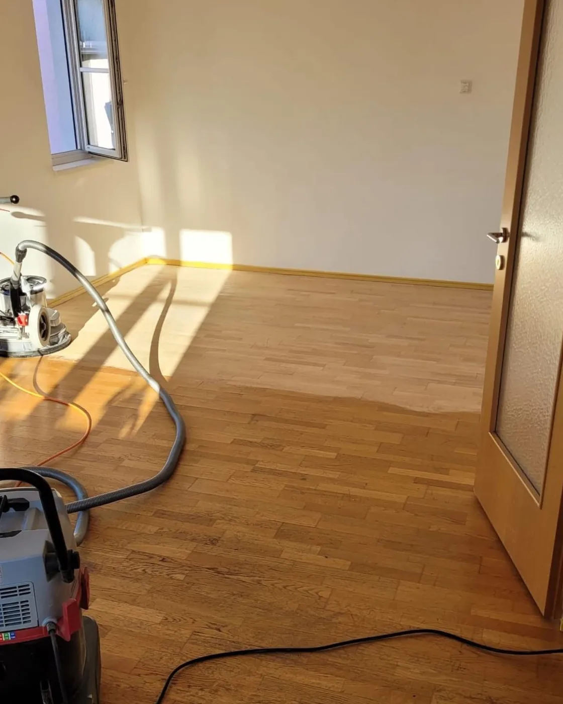 Leerer Raum mit frisch geschliffenem Holzboden, daneben Schleifmaschine und Sauger