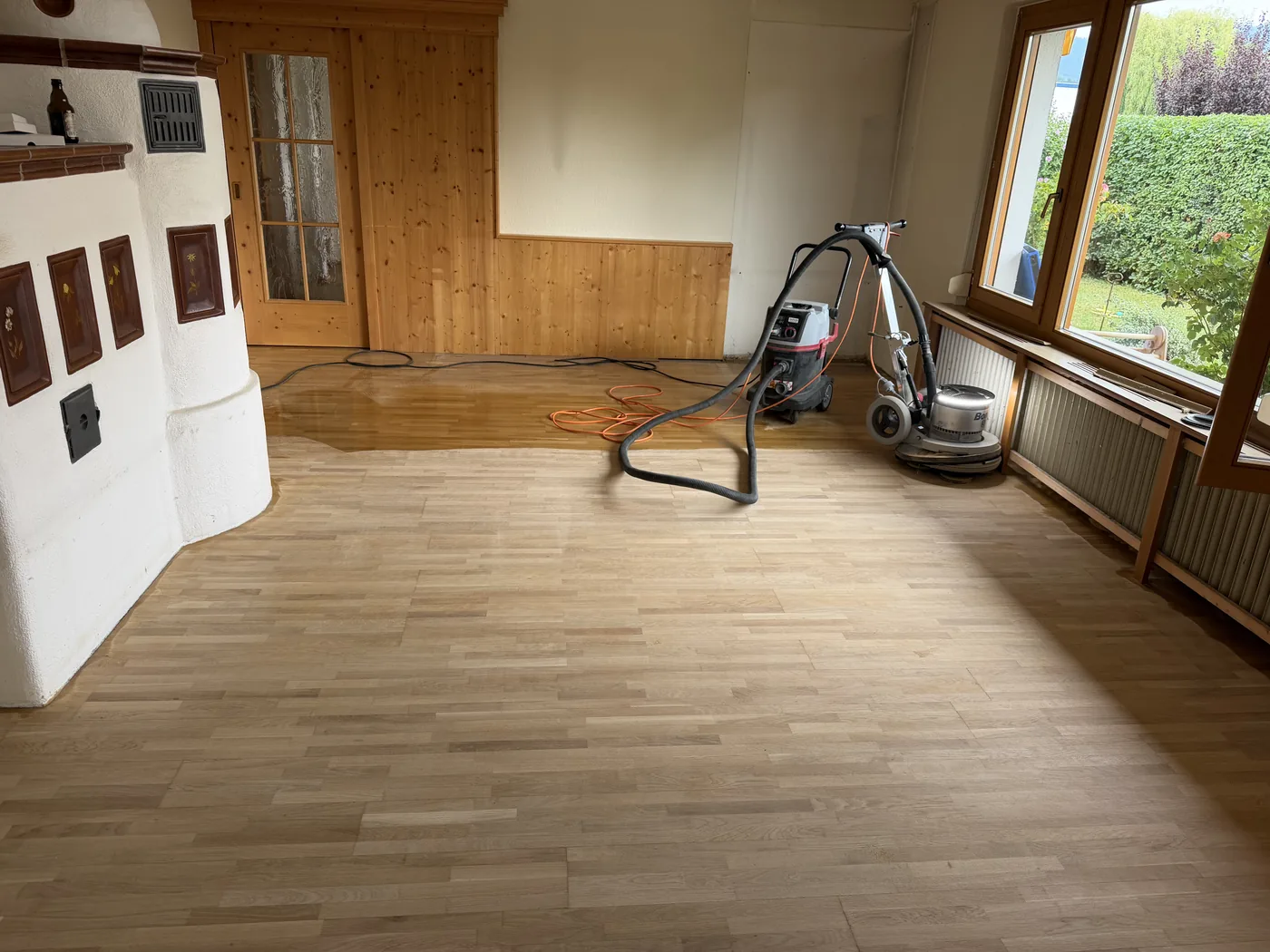 Wohnzimmer mit halb geschliffenem Holzboden, im Hintergrund die Schleifmaschine und der noch dunkle Altboden